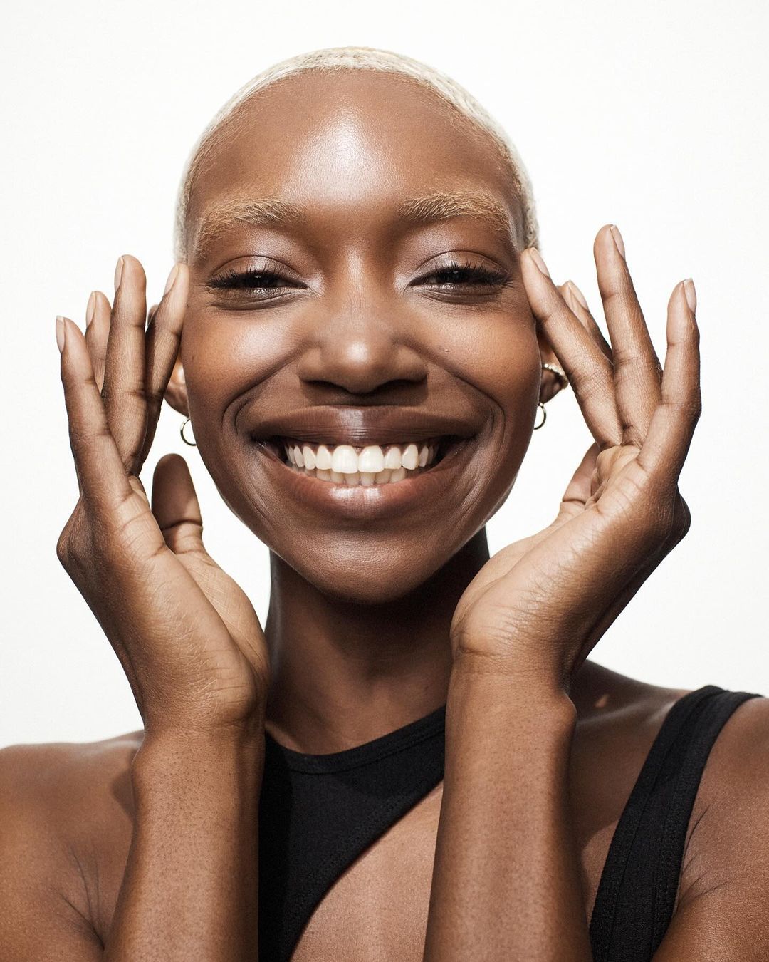Close-up studio shot of smiling Black woman with blonde hair, dark skin, black top, hands near face, and gold hoop earrings on white background.