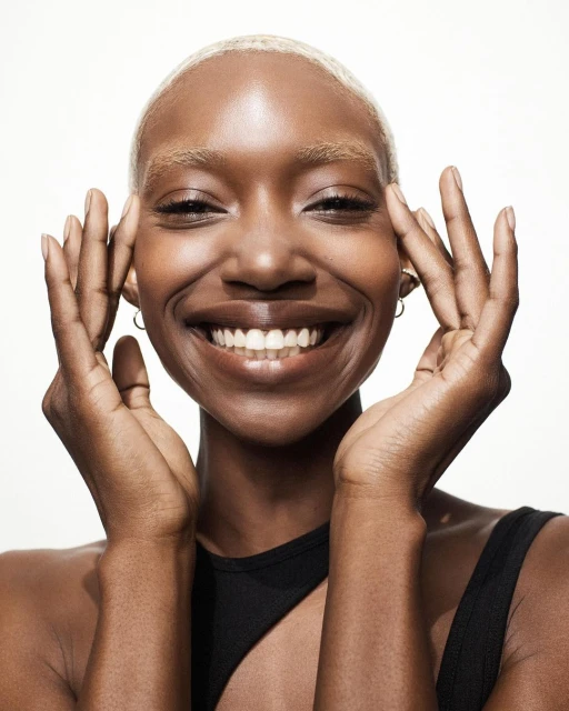 Close-up studio shot of smiling Black woman with blonde hair, dark skin, black top, hands near face, and gold hoop earrings on white background.