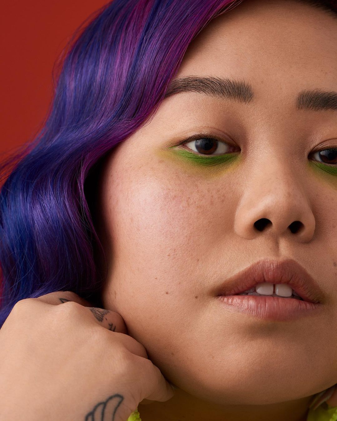 Close-up portrait of an Asian woman with green eyeshadow and dyed purple hair, posing against a red backdrop.