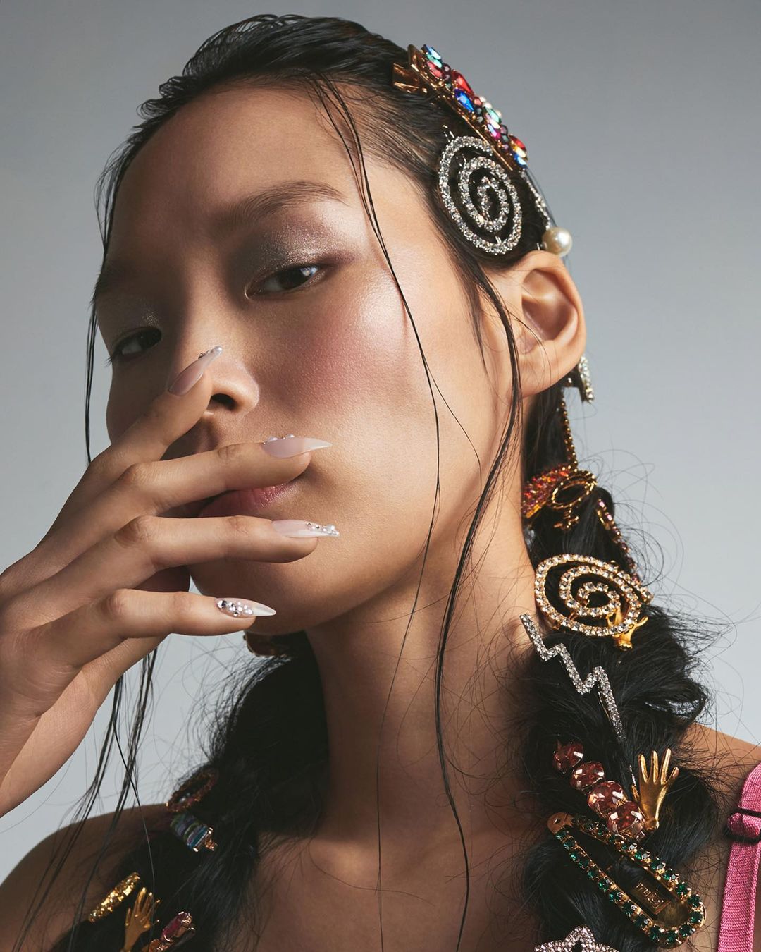Asian woman portrait with detailed makeup and jeweled hair accessories in braids posing in a studio setting with a solid light grey background.