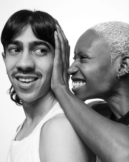 Monochrome portrait of a man smiling as a woman whispers in his ear, set against a white backdrop in a close-up studio shot.