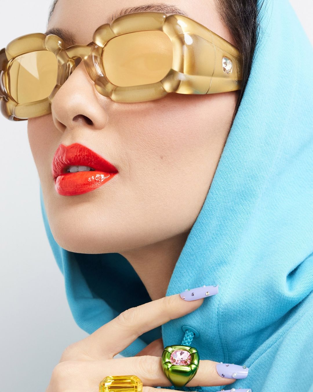 Close-up of a woman's face wearing statement sunglasses and rings, paired with a blue hoodie and light purple nail polish.