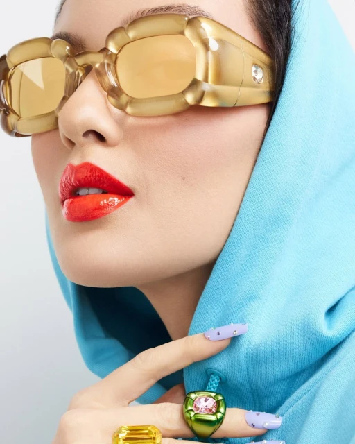 Close-up of a woman's face wearing statement sunglasses and rings, paired with a blue hoodie and light purple nail polish.