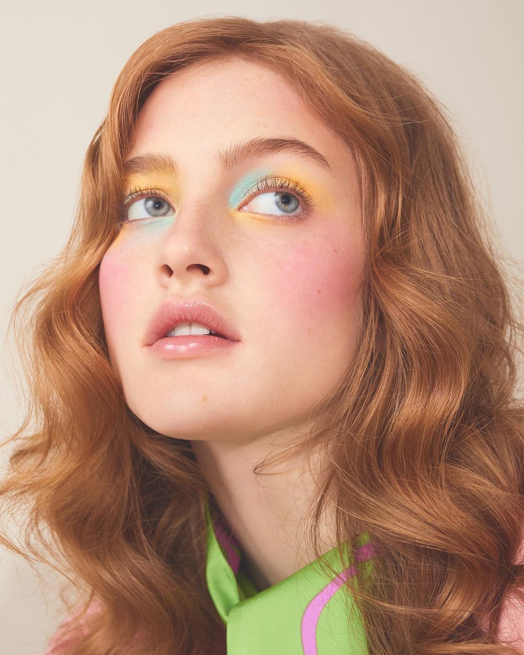 Close-up portrait of a young woman with wavy red hair, wearing colorful eye makeup, looking up against a neutral background.