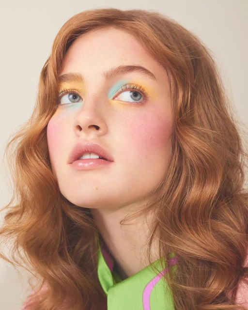 Close-up portrait of a young woman with wavy red hair, wearing colorful eye makeup, looking up against a neutral background.