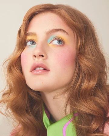Close-up portrait of a young woman with wavy red hair, wearing colorful eye makeup, looking up against a neutral background.