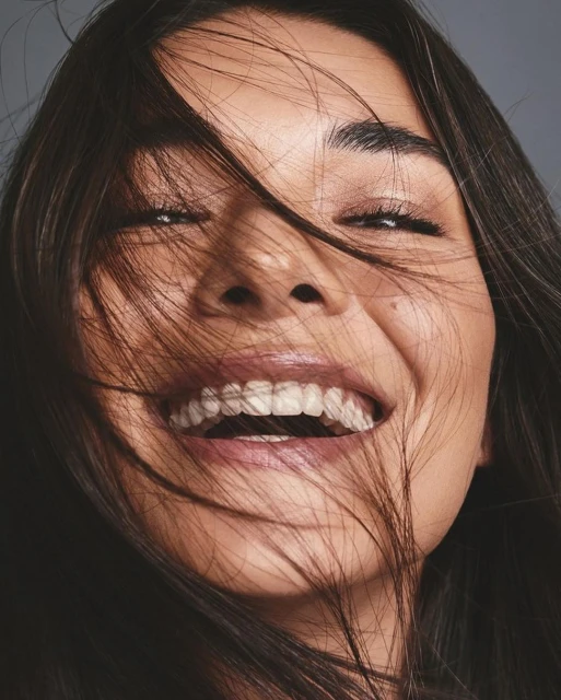 Close-up of a woman with dark hair smiling broadly, with strands of hair across her face, showcasing a joyful and candid moment.