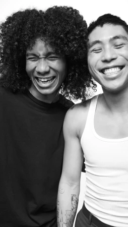 Two young adults smile brightly in a black and white portrait. One has curly hair and a black shirt; the other wears a white tank top.