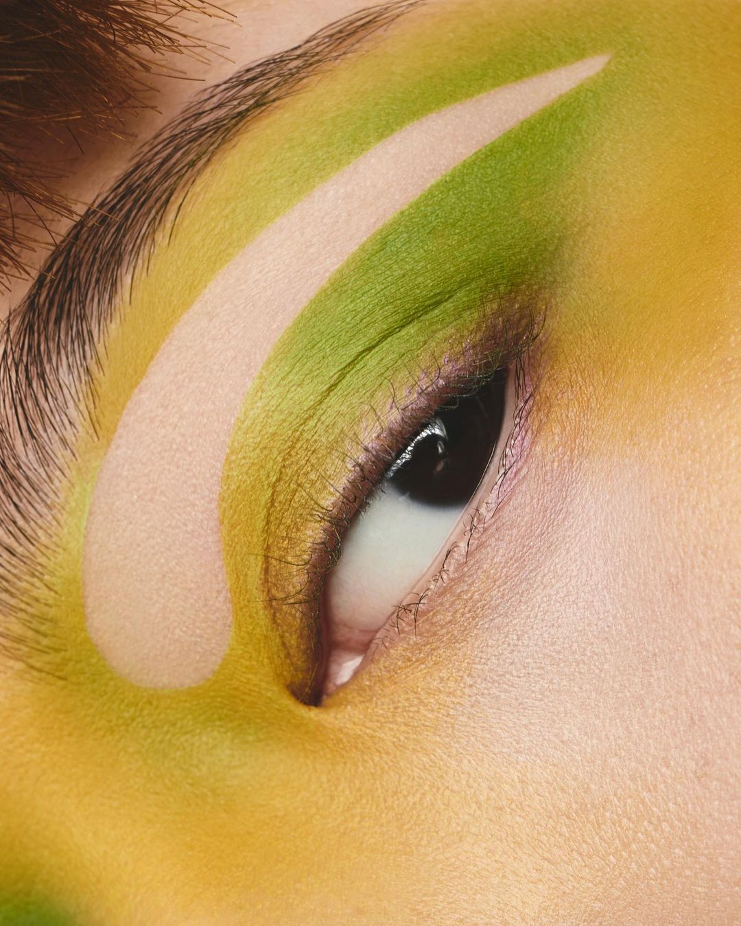 Close-up on an eye with green and yellow eyeshadow. The graphic makeup includes a thick white stripe above the crease.