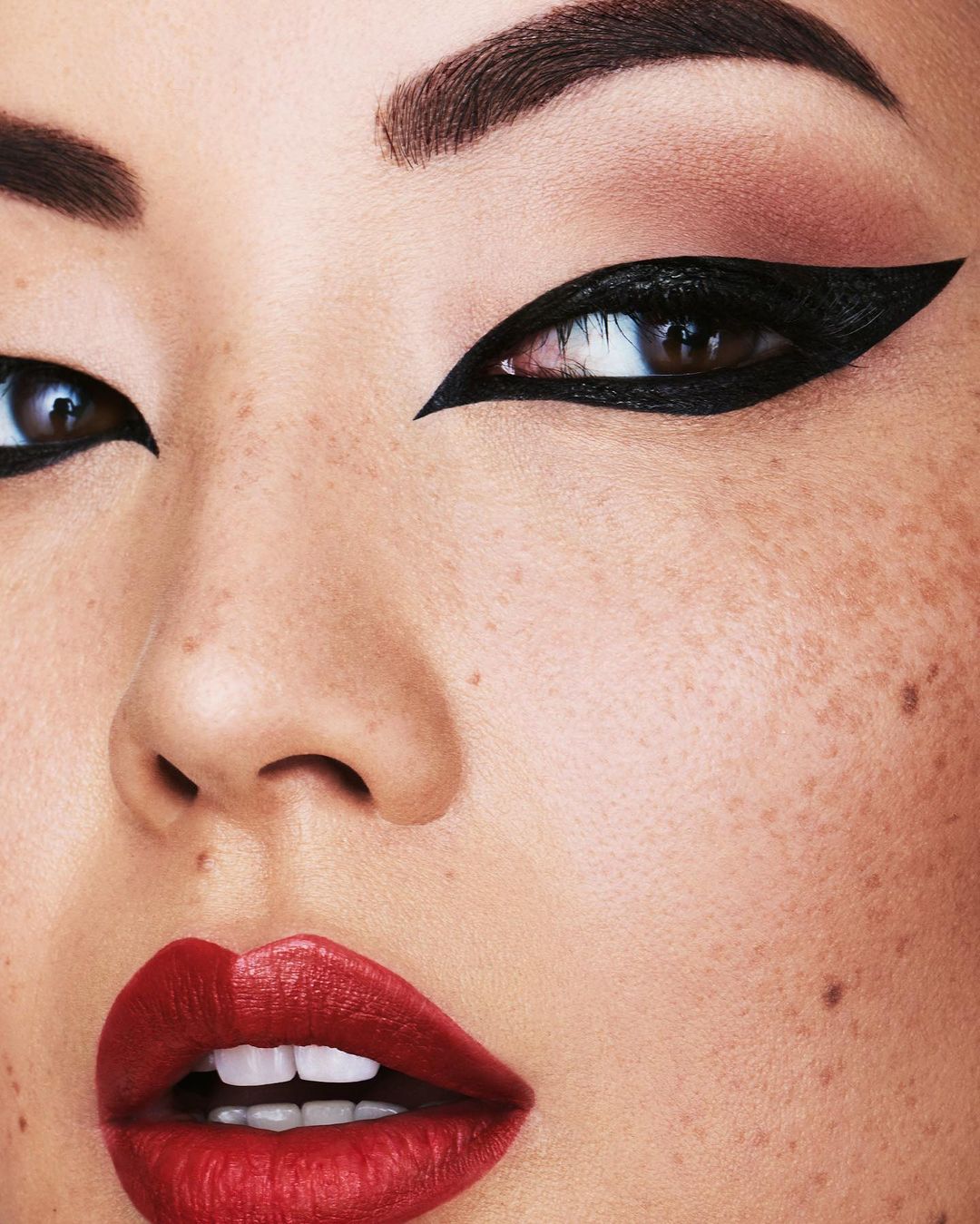 Close-up of an Asian woman's face showcasing striking black eyeliner, bold red lipstick, and prominent freckles. Beauty and fashion makeup.