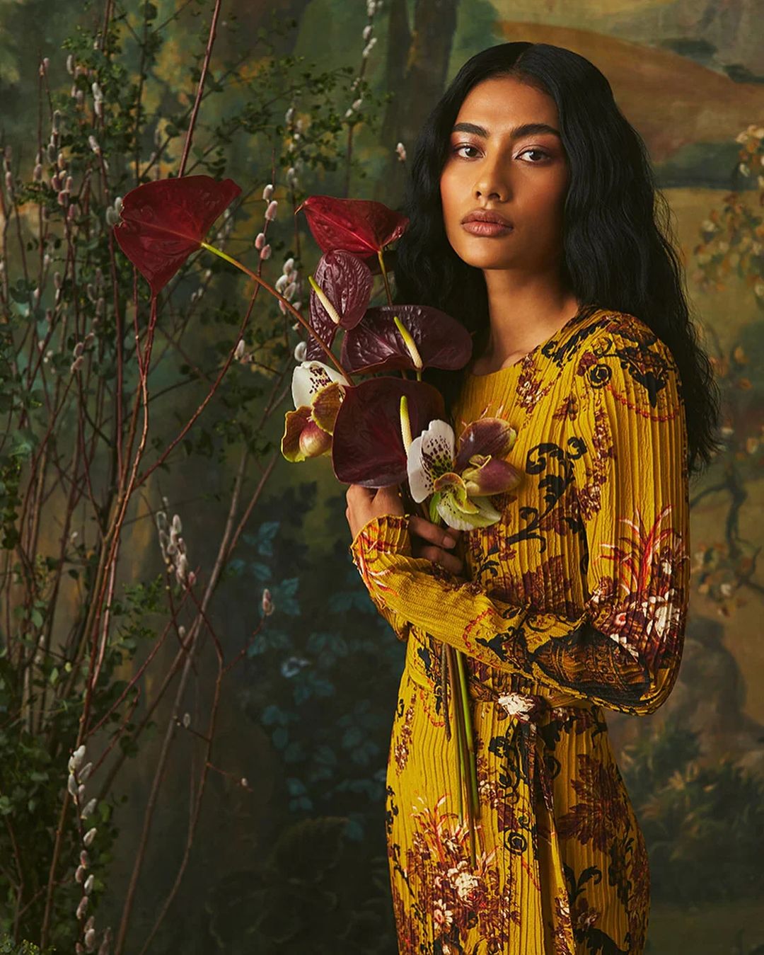 Portrait of an adult woman with long dark hair holding red Anthurium flowers while posing in front of a painterly, floral backdrop.