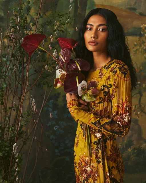Portrait of an adult woman with long dark hair holding red Anthurium flowers while posing in front of a painterly, floral backdrop.