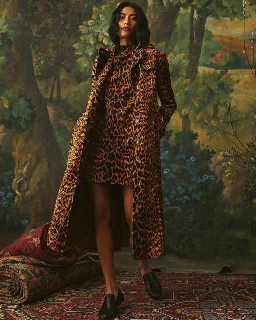 A woman poses in a full-length leopard print trench coat and matching dress in a painted forest scene, complemented by patterned rugs.