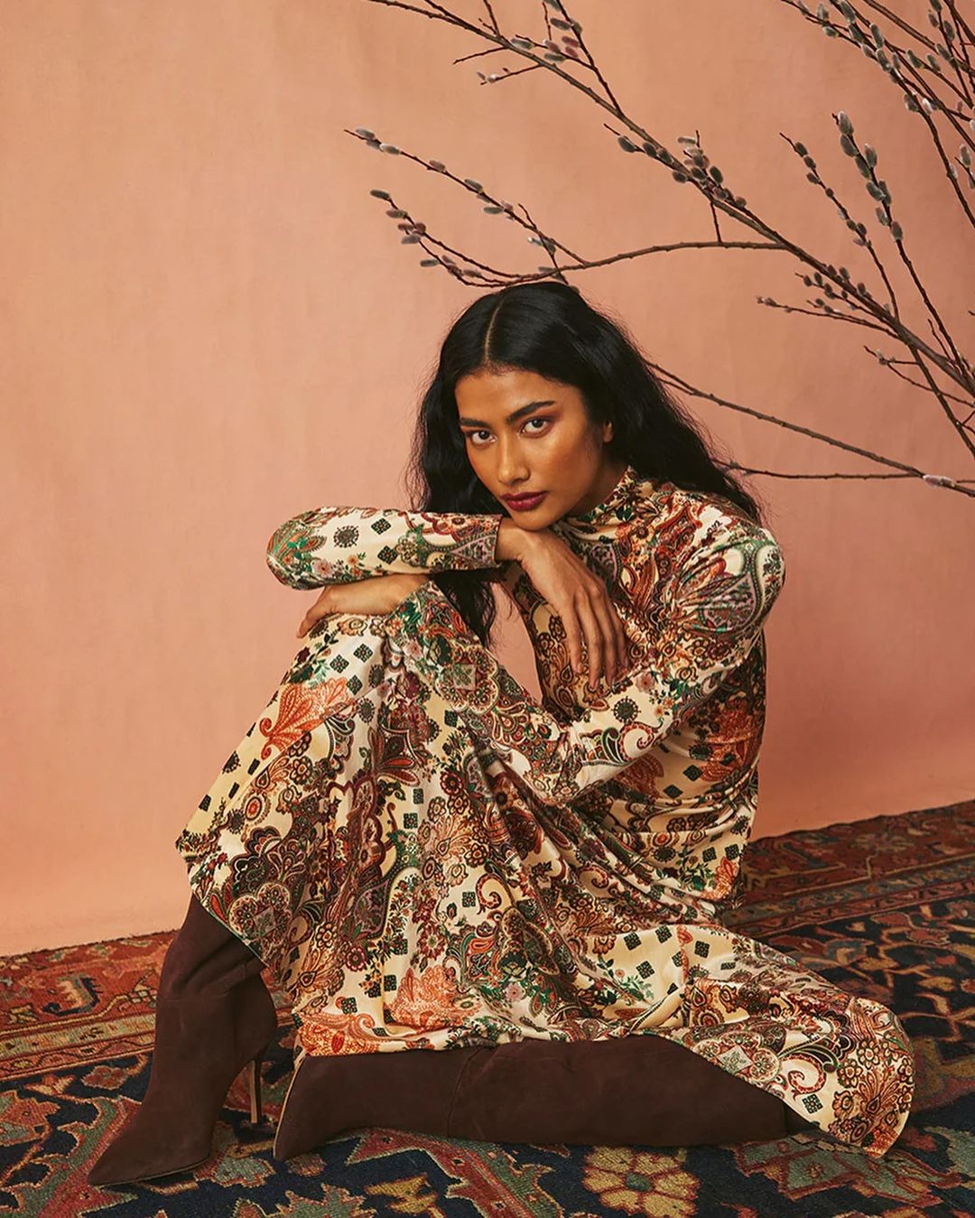 A woman in a patterned dress and boots poses on a rug in front of a peach-colored wall with a bare branch, looking at the camera.