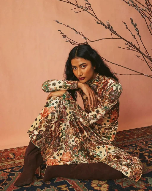 A woman in a patterned dress and boots poses on a rug in front of a peach-colored wall with a bare branch, looking at the camera.