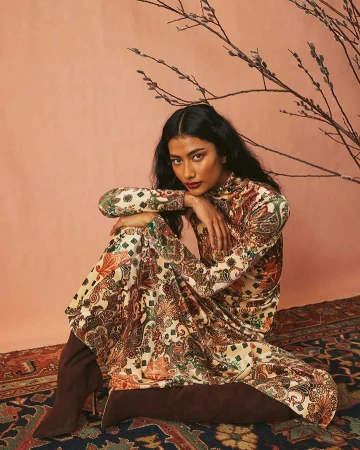 A woman in a patterned dress and boots poses on a rug in front of a peach-colored wall with a bare branch, looking at the camera.
