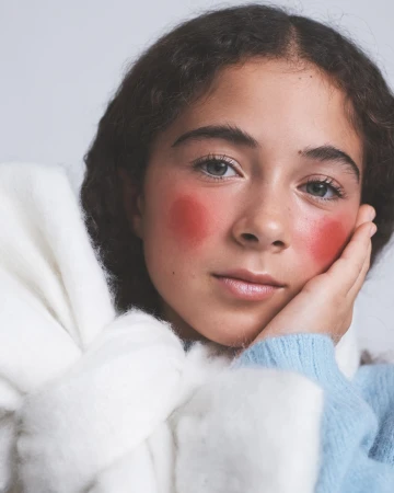 Close-up portrait of a young woman with pink blush makeup resting her hand on her cheek against a plain white background.