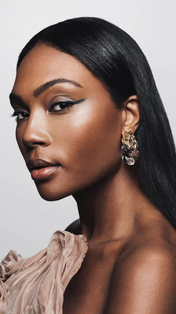 Close-up of an adult African American woman in a strapless top with winged eyeliner and gold statement earrings against a white backdrop.