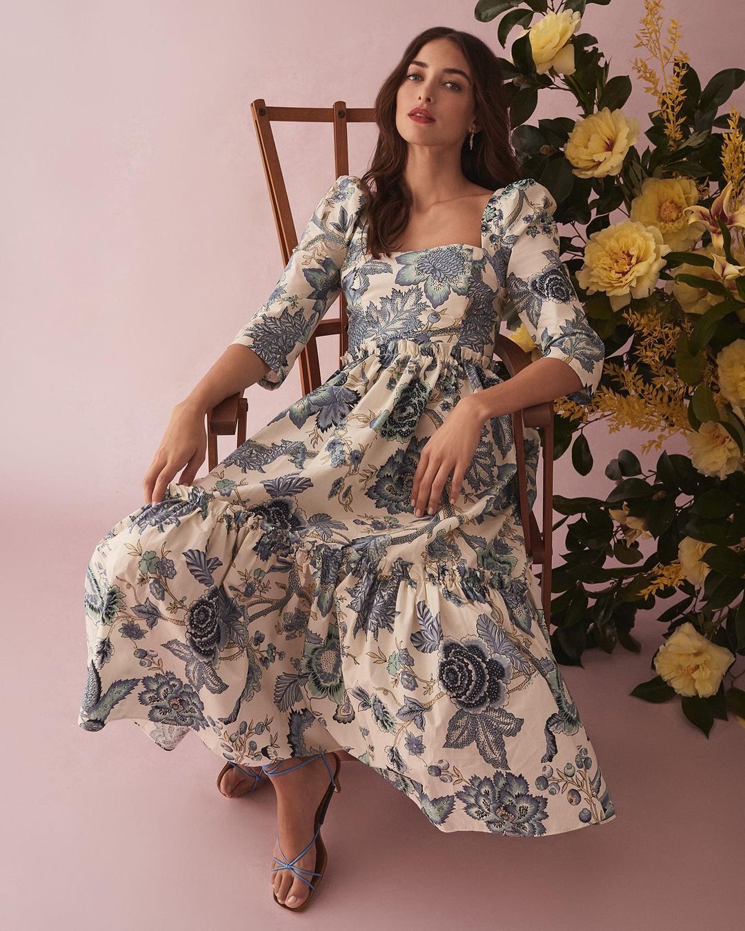 Woman in floral dress seated next to yellow flowers. The dress has blue floral designs on an off-white fabric in a studio setting.