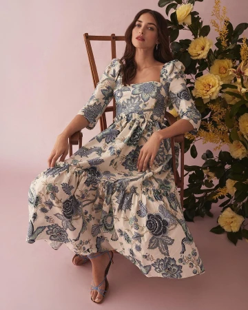 Woman in floral dress seated next to yellow flowers. The dress has blue floral designs on an off-white fabric in a studio setting.