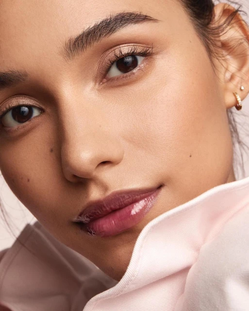 Close-up of a young woman's face, featuring glossy lips, natural makeup, and a pink collared shirt against a soft, neutral background.