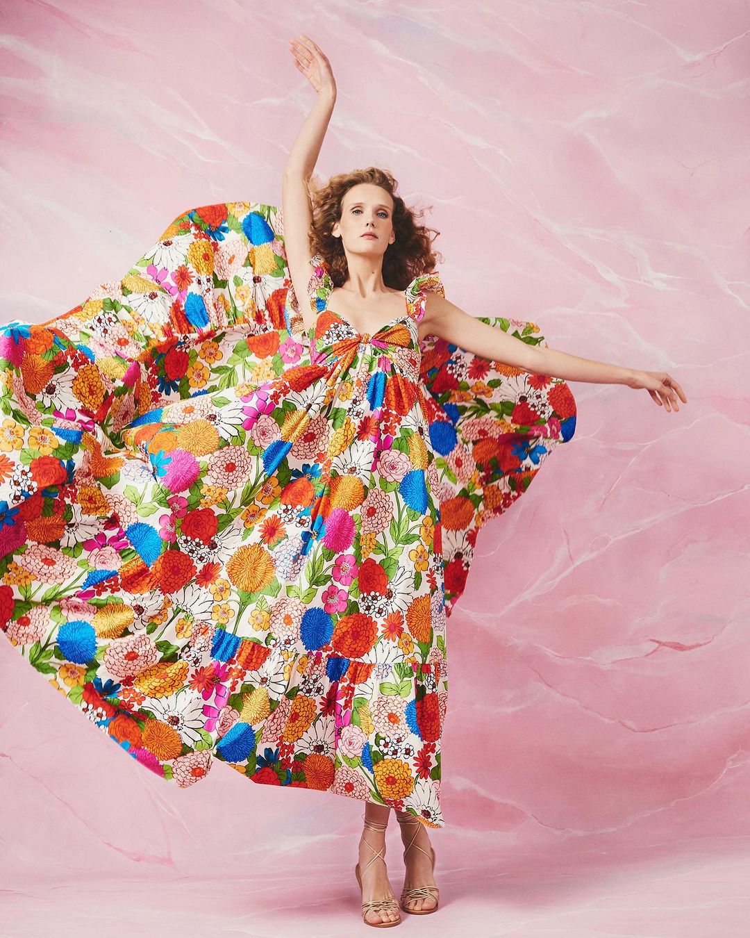 Adult female model wearing a flowing white dress with a multicolored floral print in a photo studio with a pink marble backdrop.