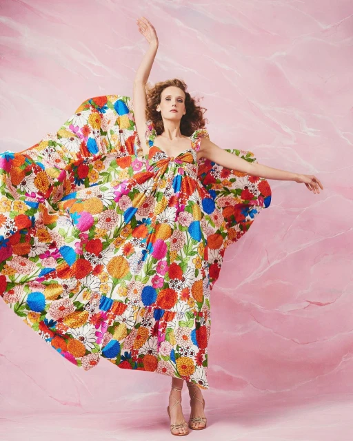 Adult female model wearing a flowing white dress with a multicolored floral print in a photo studio with a pink marble backdrop.