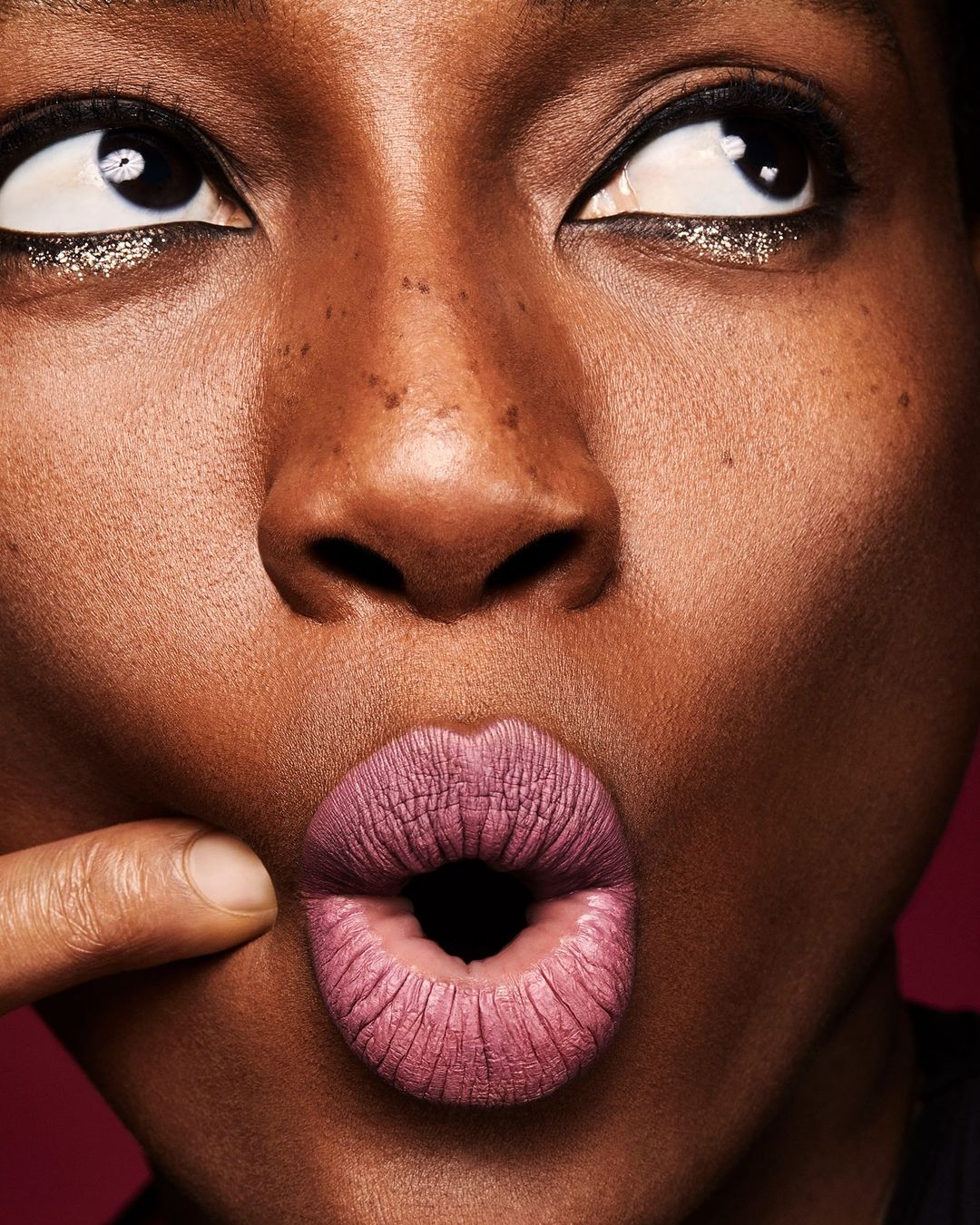 Close-up of an African-American female's face, featuring her dark skin, freckles, wide eyes, and a finger pulling her mauve-colored lips.