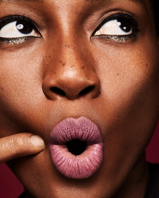 Close-up of an African-American female's face, featuring her dark skin, freckles, wide eyes, and a finger pulling her mauve-colored lips.