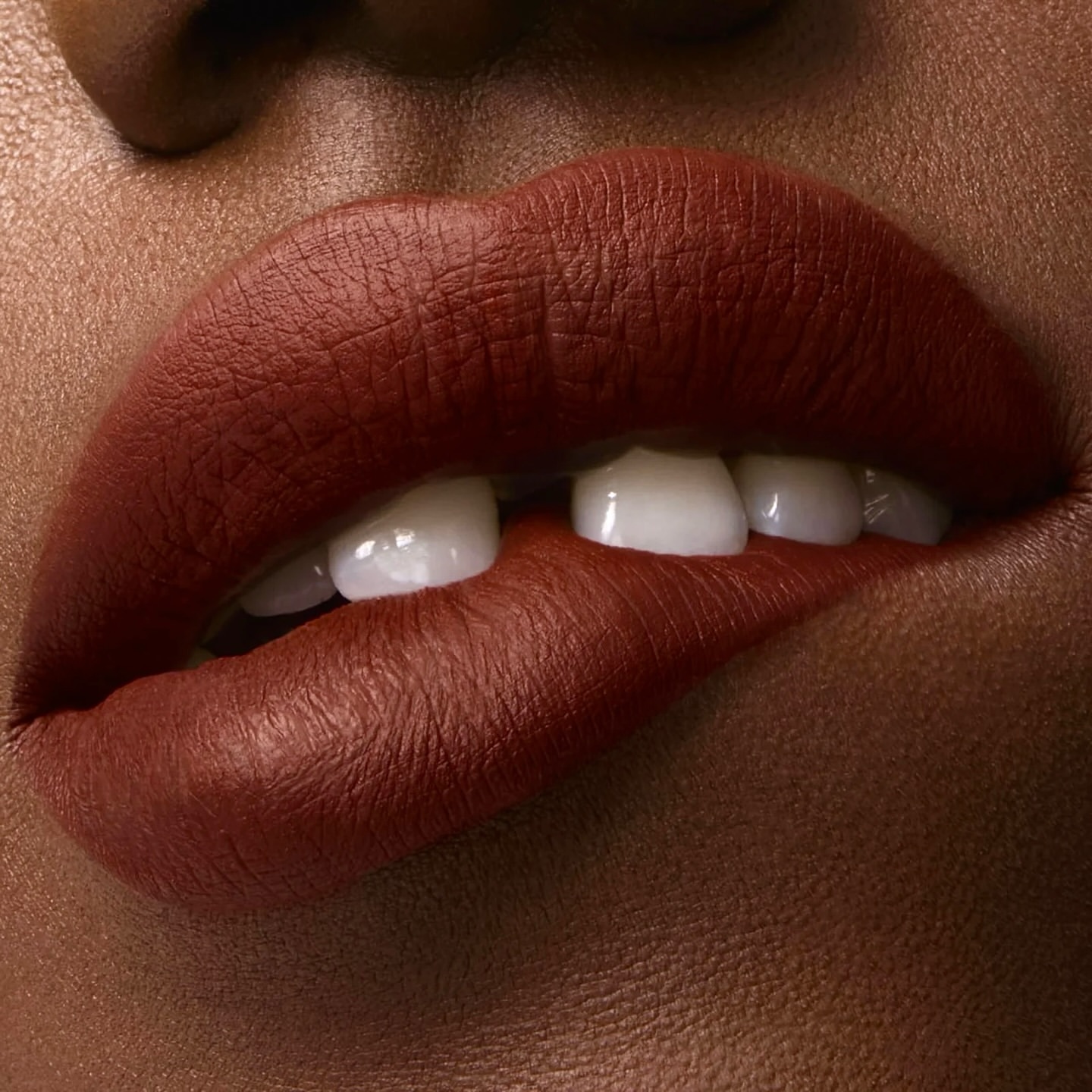 Close-up shot of an adult female with dark skin biting her lower lip, displaying a bold red matte lipstick and white teeth.