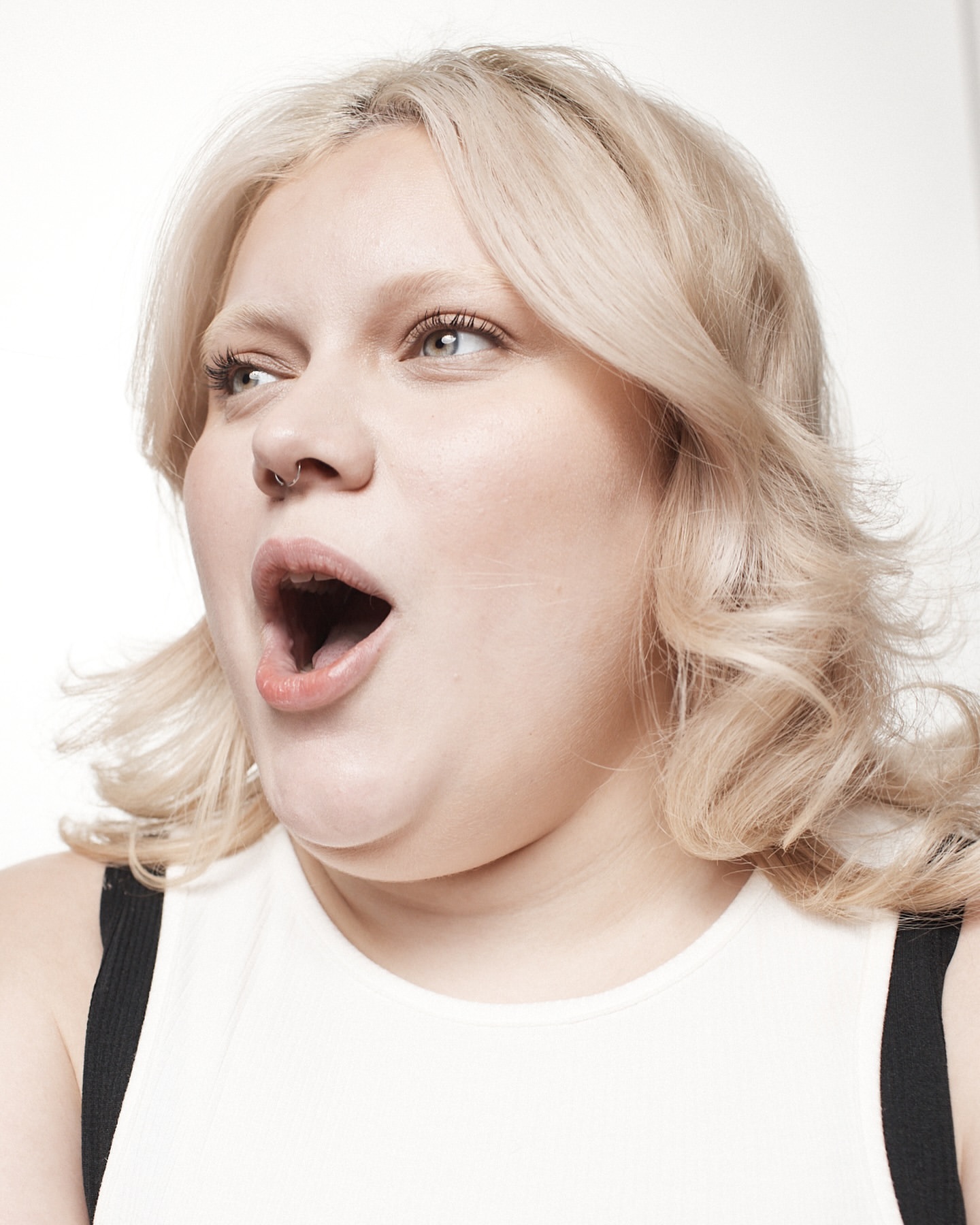 Portrait of a blonde adult female with an open mouth, wearing a white top. Indoor studio shot against a bright white background.