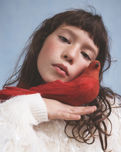 Close-up of a child model holding a bright red bird, wearing a textured white sweater, against a light blue backdrop in a studio setting.