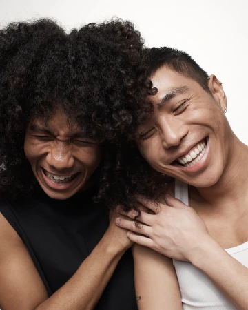 Two smiling men with close-cropped and curly hair posing together against a plain white background