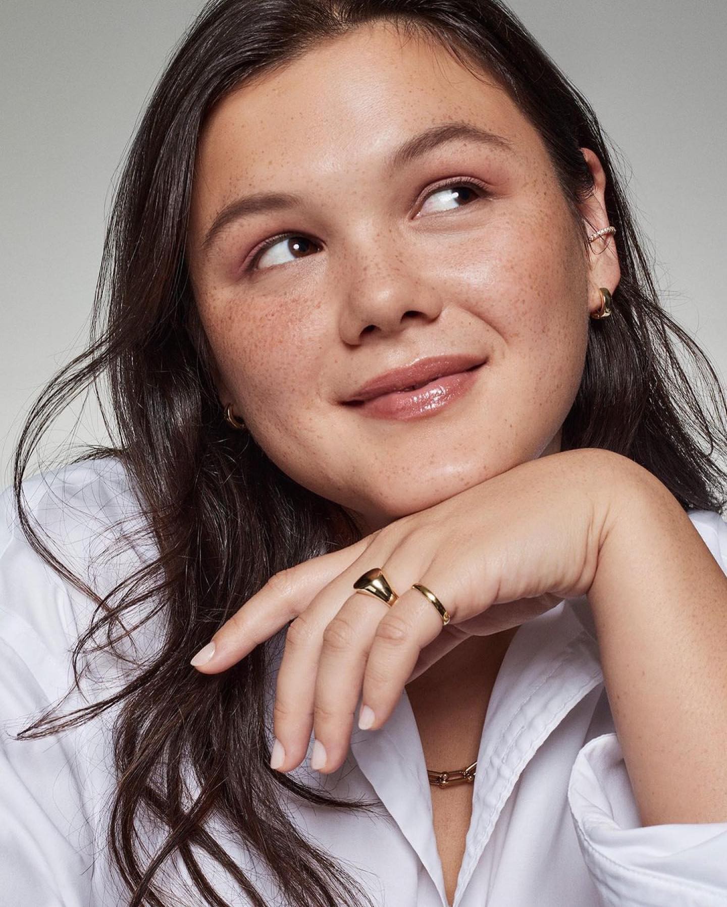 Close up portrait of a young woman resting her chin on her hand, wearing a white shirt and gold rings against a neutral background.