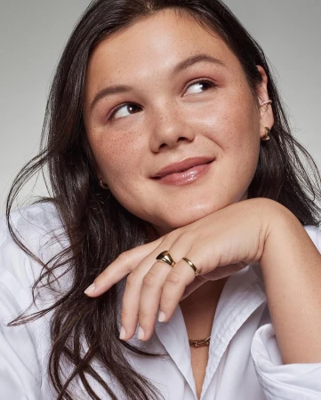 Close up portrait of a young woman resting her chin on her hand, wearing a white shirt and gold rings against a neutral background.
