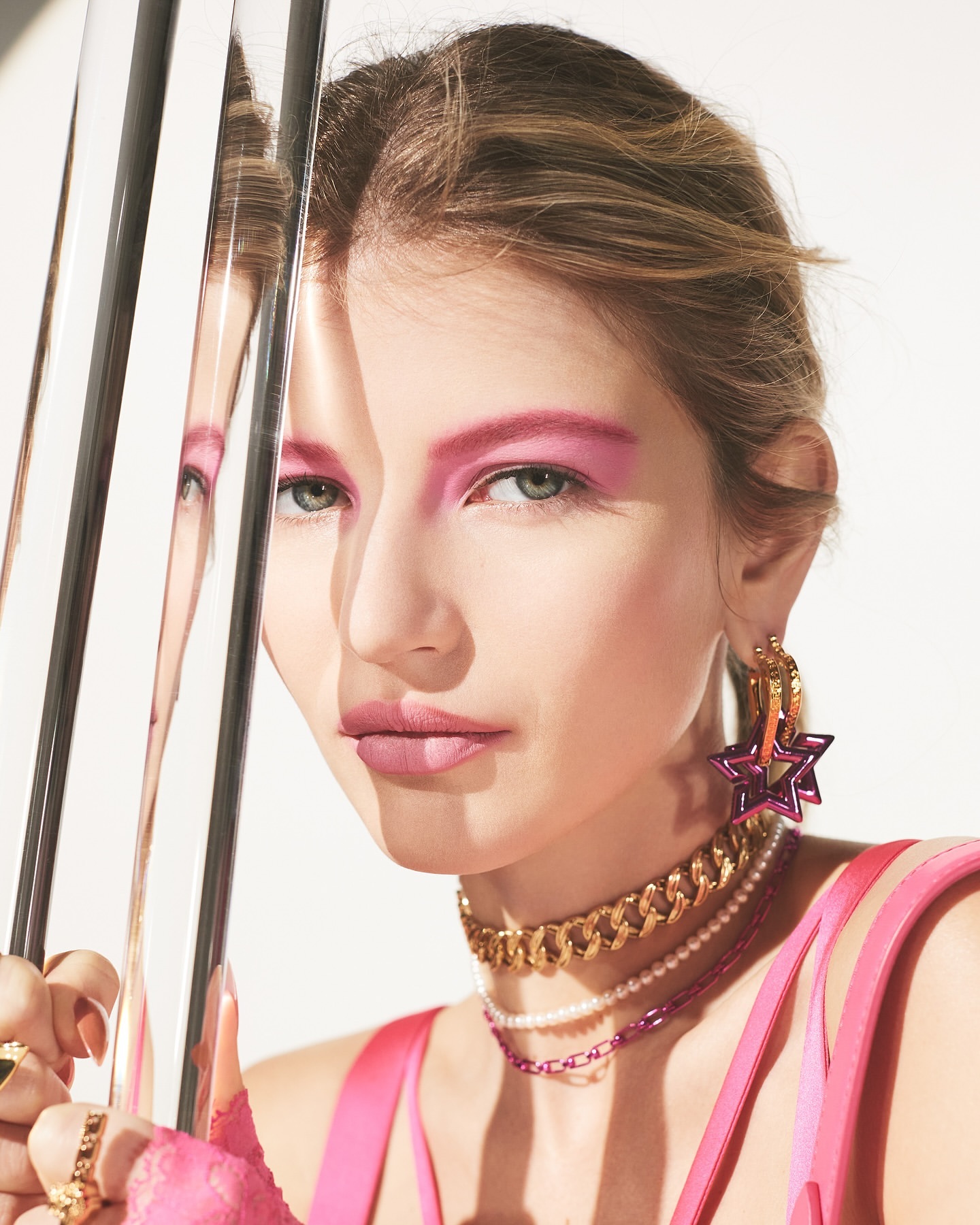 Close-up of a fair-skinned model with bold pink makeup, gold jewelry, and layered necklaces, reflecting in metallic bars on a white background.