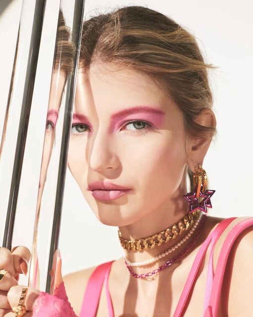 Close-up of a fair-skinned model with bold pink makeup, gold jewelry, and layered necklaces, reflecting in metallic bars on a white background.