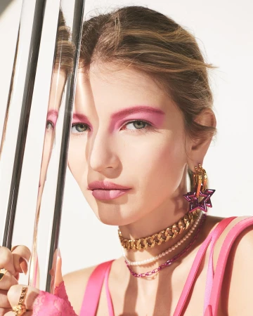 Close-up of a fair-skinned model with bold pink makeup, gold jewelry, and layered necklaces, reflecting in metallic bars on a white background.