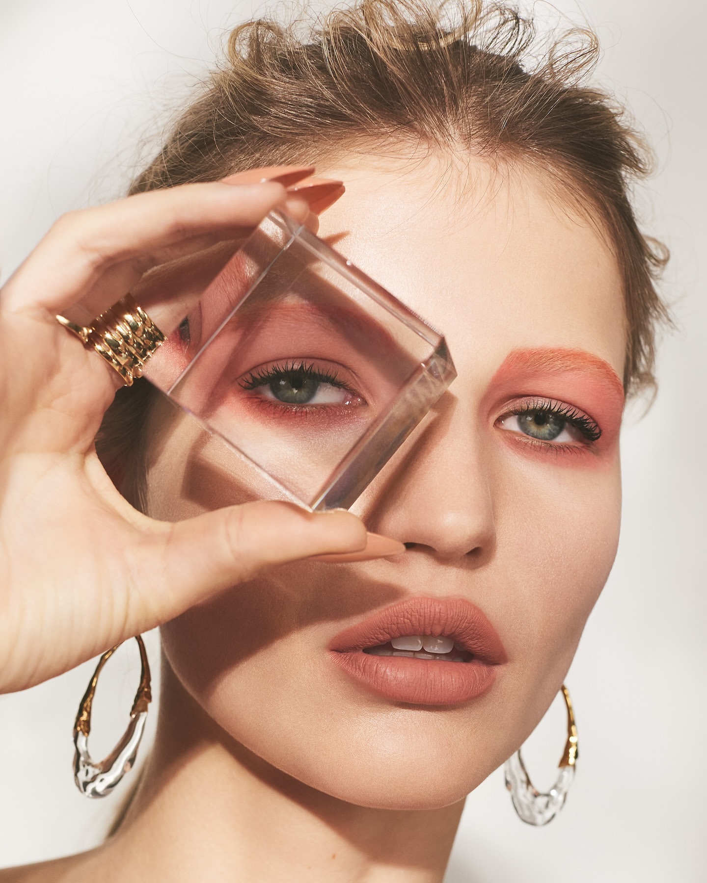 Close-up of a woman with fair skin, artistic peach-toned makeup, and sculptural gold earrings, holding a clear cube.