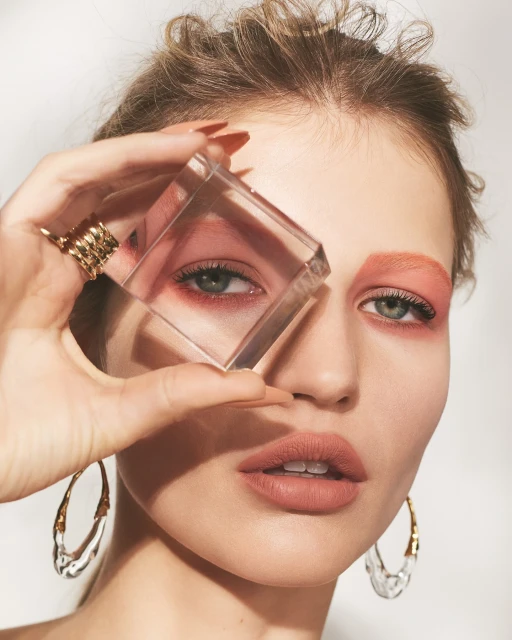 Close-up of a woman with fair skin, artistic peach-toned makeup, and sculptural gold earrings, holding a clear cube.