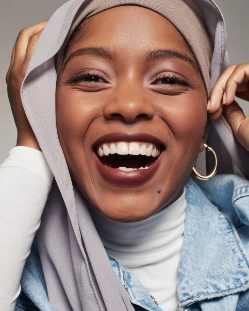 Close-up of a smiling African-American woman wearing a light-grey hijab, denim jacket, and gold hoop earring against a neutral backdrop.