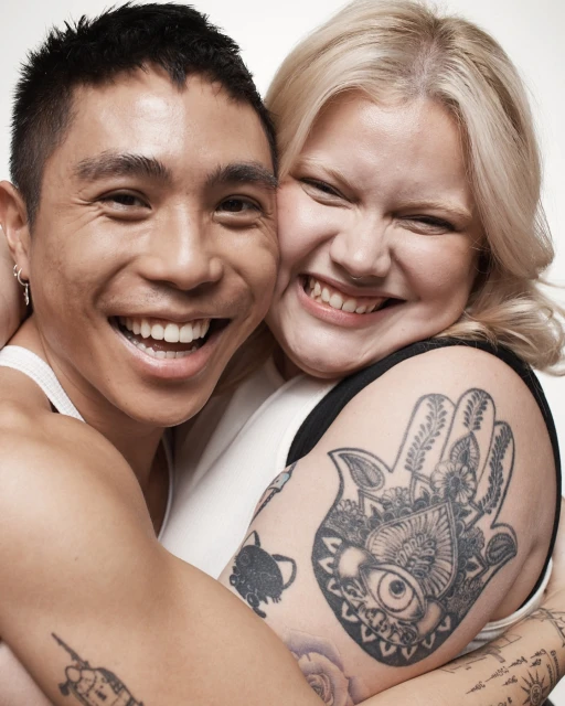 Close-up studio portrait of a happy, smiling mixed-race couple hugging tightly, with tattoos visible on their arms against a white backdrop.