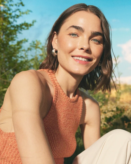 Smiling brunette woman with silver hoop earrings posing in a sleeveless, orange knit top, outdoors with blurred trees and blue sky.