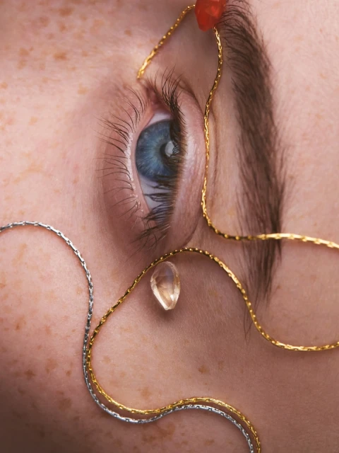 Close-up portrait of a person's eye with silver and gold chain jewelry, freckled skin, and a blue iris looking at the camera.