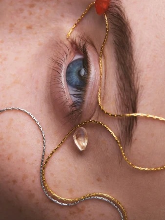 Close-up portrait of a person's eye with silver and gold chain jewelry, freckled skin, and a blue iris looking at the camera.
