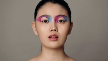 Portrait of a woman with colorful makeup on her upper eyelids, set against a neutral grey background in a studio setting.