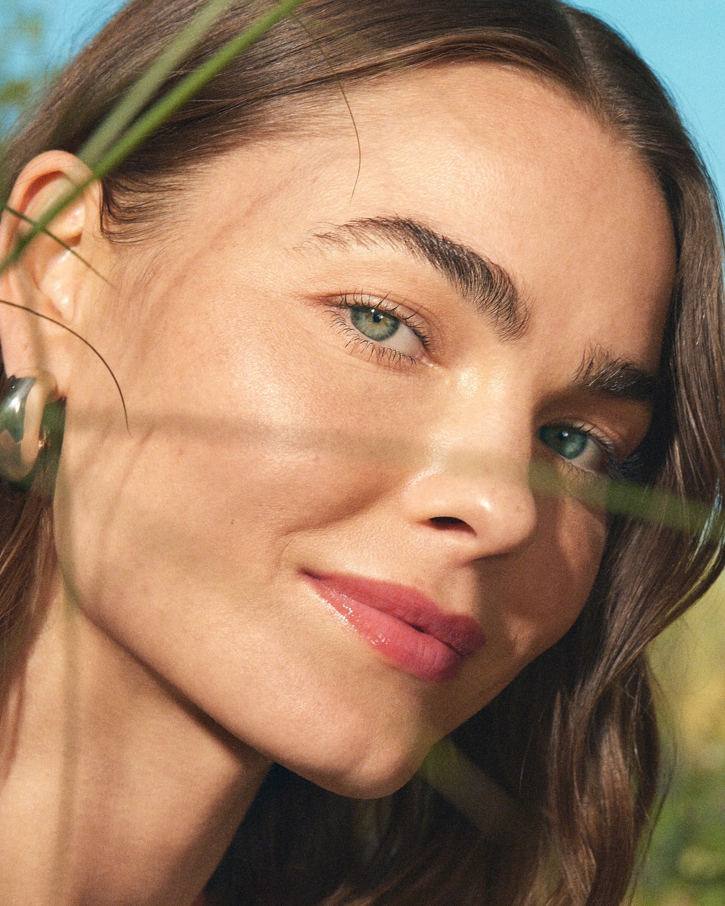 Close-up of an adult female with brown hair and light eyes, looking directly at the camera with a slight smile, and green grass.
