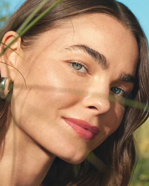 Close-up of an adult female with brown hair and light eyes, looking directly at the camera with a slight smile, and green grass.