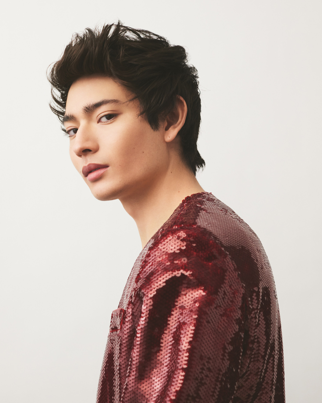 Portrait of a young adult man with dark hair, fair skin, wearing a sequined burgundy long-sleeved shirt, posing against a white background.