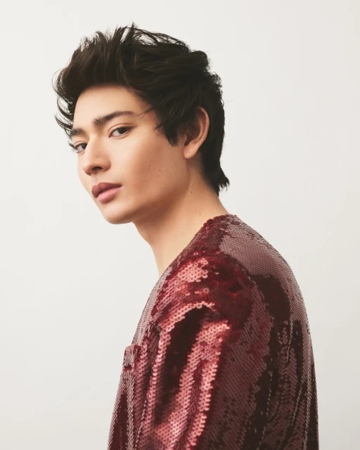 Portrait of a young adult man with dark hair, fair skin, wearing a sequined burgundy long-sleeved shirt, posing against a white background.